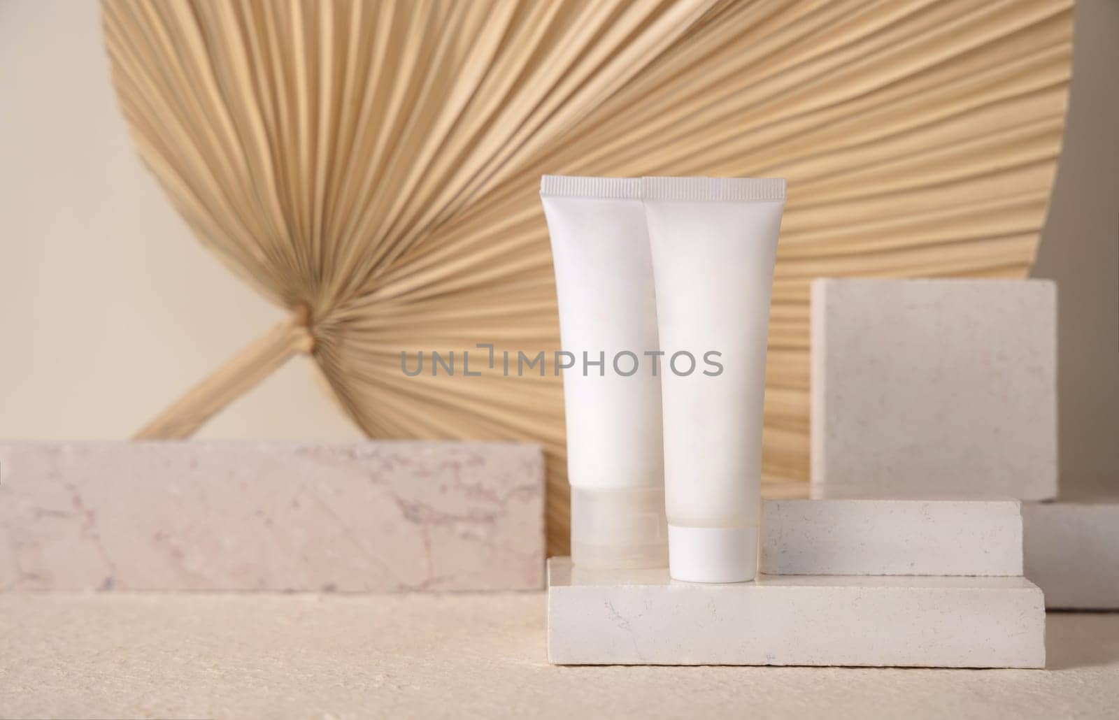 White blank tubes on travertine stone against dry beige palm leaf close up, mockup. Skincare beauty product - cream or lotion. Natural organic cosmetics. Minimal eco friendly composition..