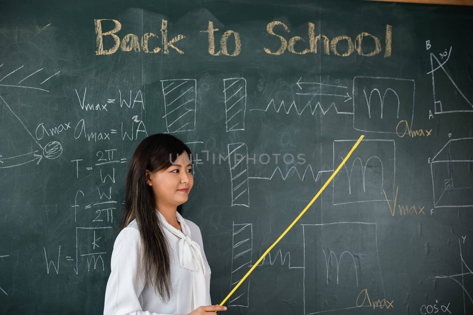 Back to school concept. Asian female teacher smiling with wooden stick pointing to blackboard at school in classroom, Happy beautiful young woman standing hold pointer to back board