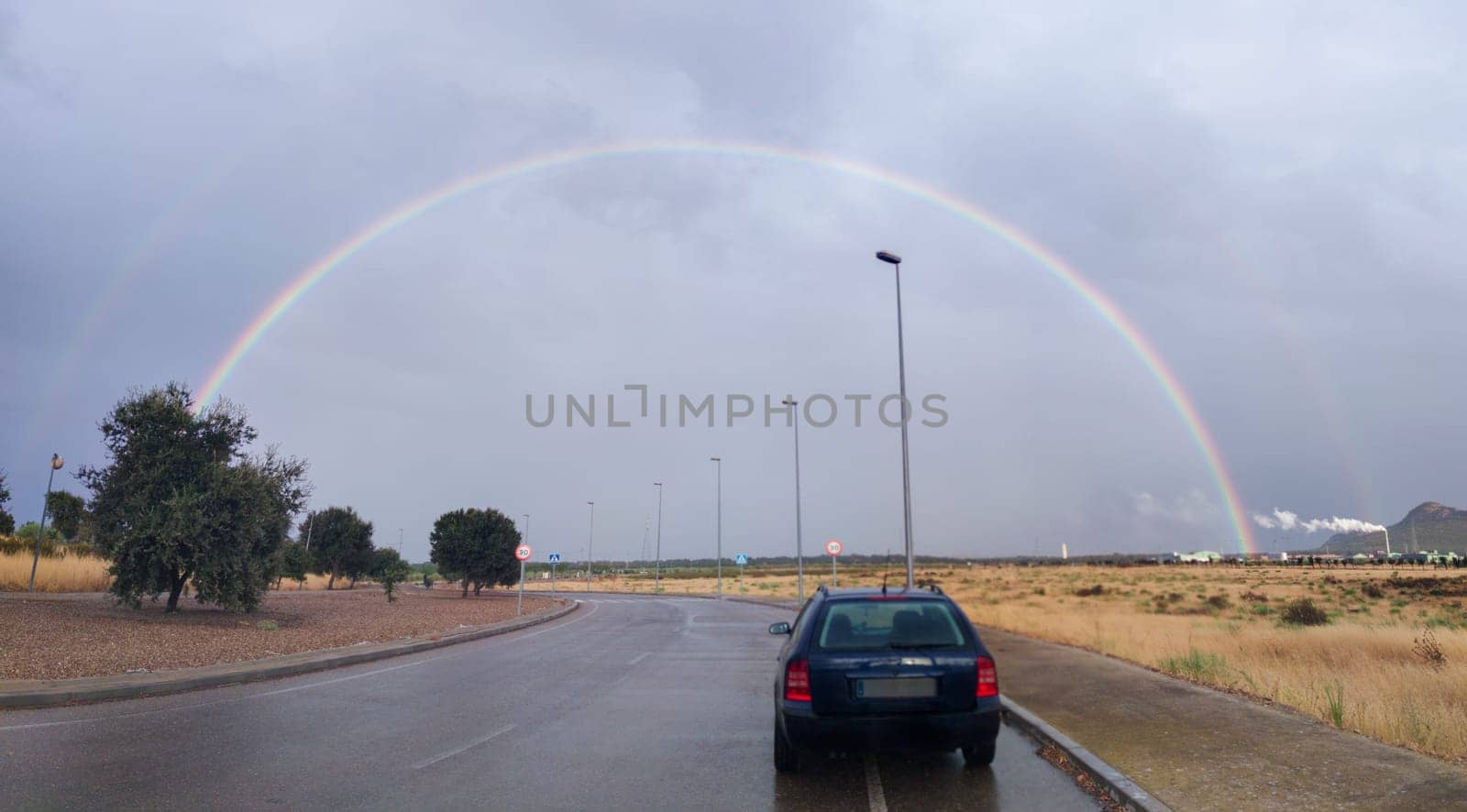 Car at roundabout with rainbow rising over horizon. Industrial area
