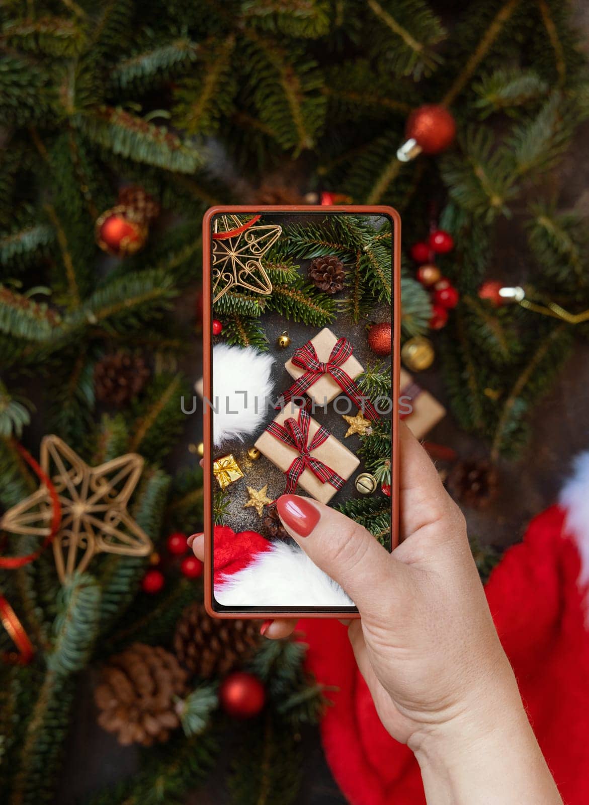 Woman's hand taking a mobile photo of christmas composition with Santa Claus hat, fir branches and holiday decorations top view. Blogger preparing content for social media using cell phone.