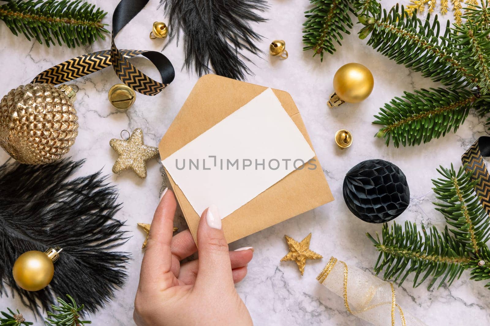 Hand holding Card and envelope, black and golden Christmas decorations and fir tree branches, top view. Winter flat lay with blank invitation card, Mockup, copy space