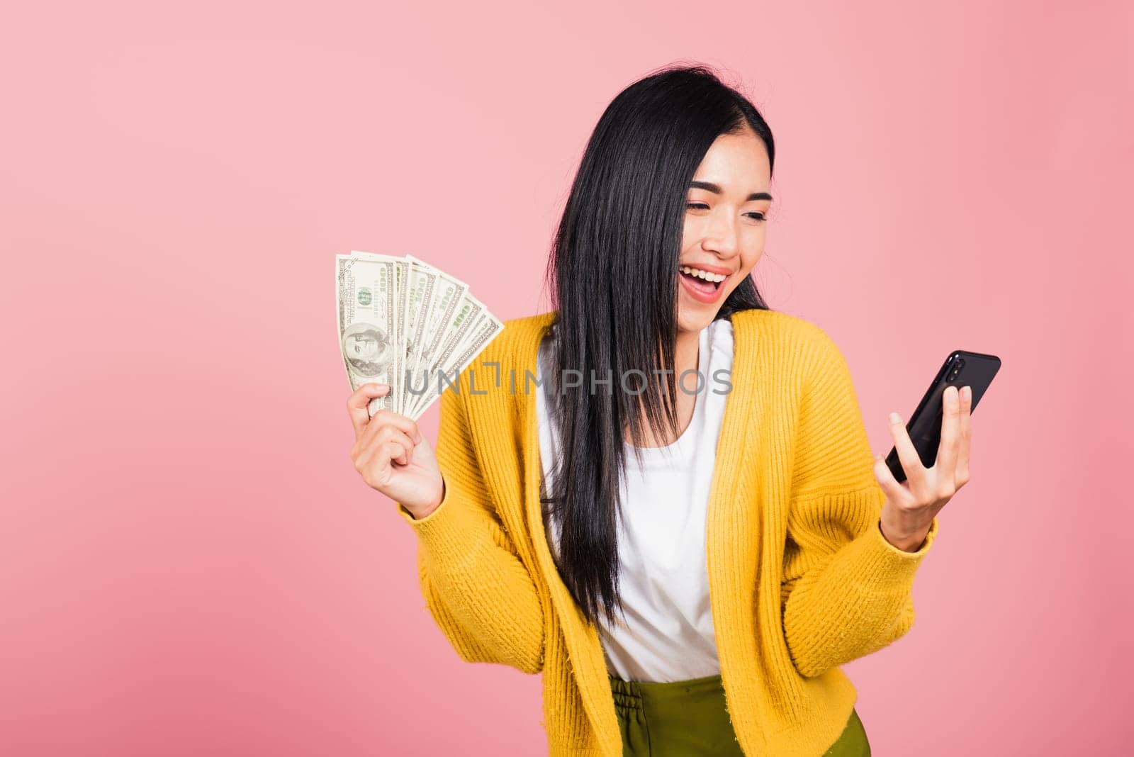 Portrait Asian happy beautiful young woman teen shopper smiling standing excited holding online smart mobile phone and dollar money banknotes on hand in summer, studio shot isolated on pink background