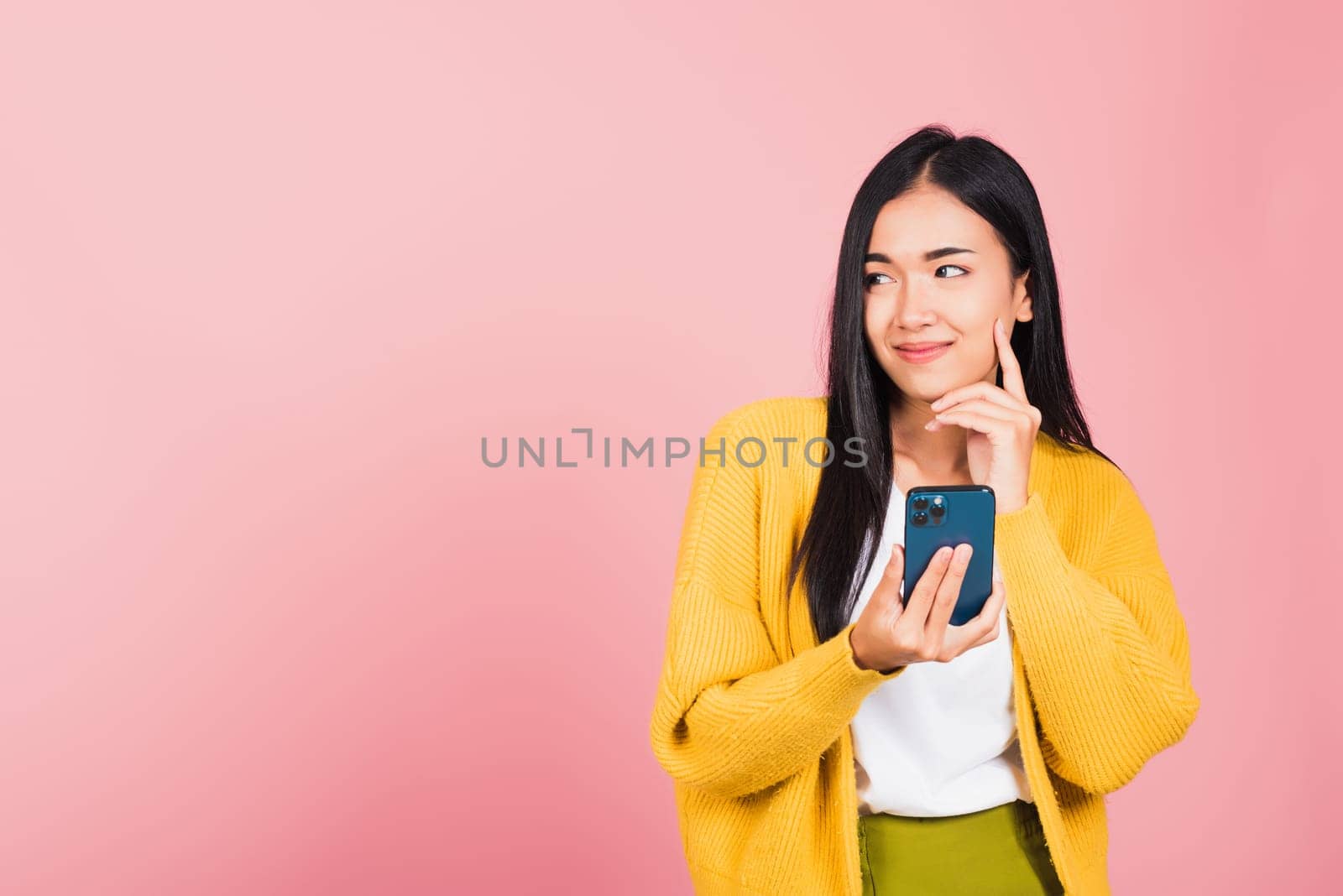 Happy Asian portrait beautiful cute young woman excited smiling holding mobile phone and think idea finger touch face, studio shot isolated on pink background, female using smartphone making gesture