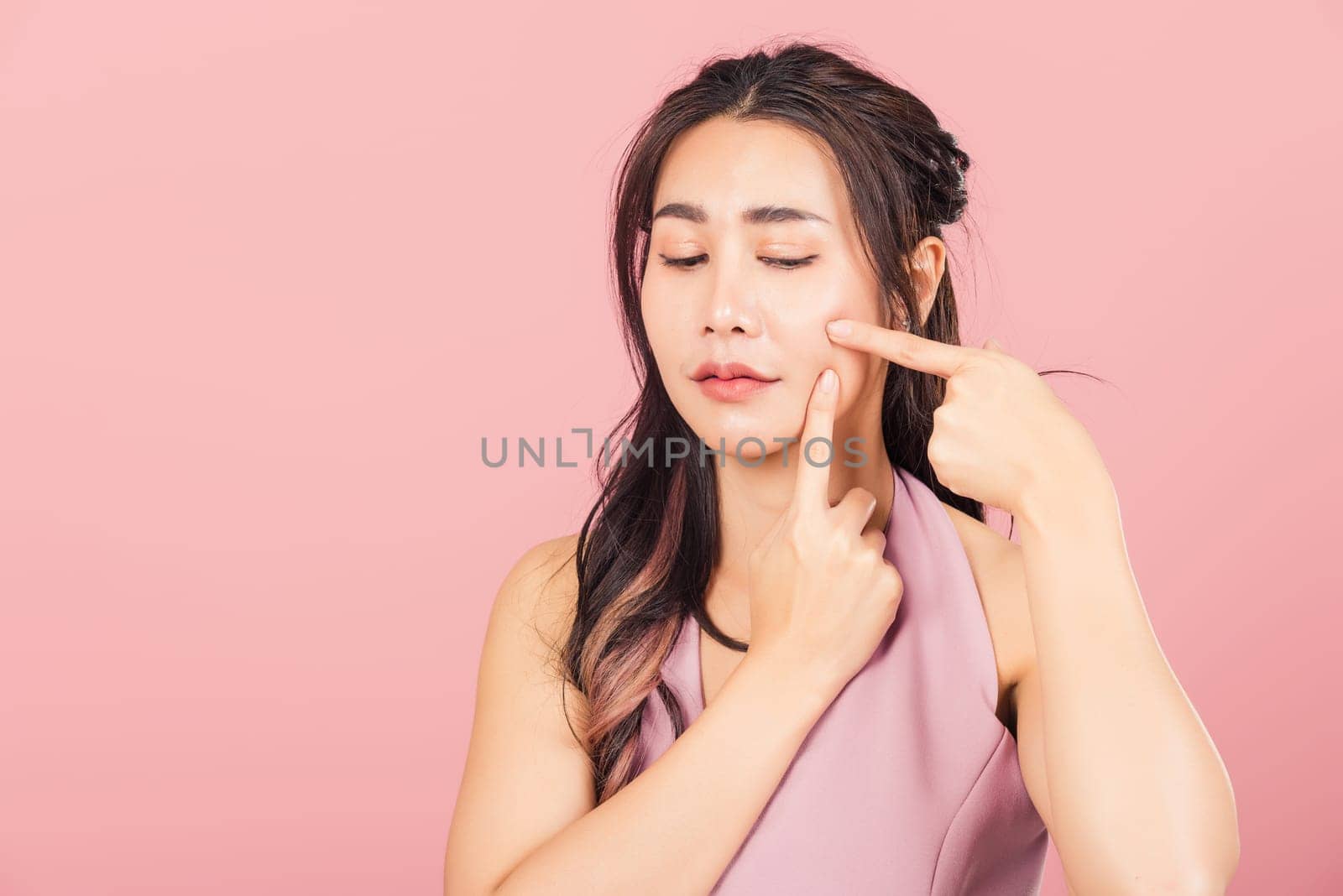 Portrait teenage Asian beautiful young woman having skin problems squeezing pimples on her face, studio shot on pink background, with copy space, Thai female acne, beauty care concept