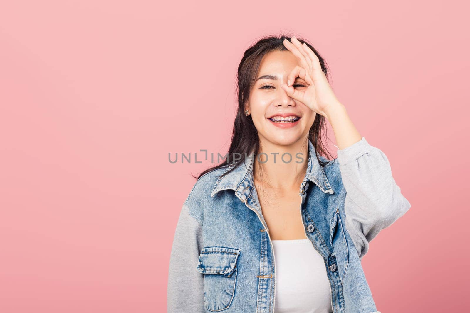 Portrait Asian beautiful young woman standing, She made finger OK symbol sign to agree near eyes looking at camera, studio shot isolated on pink background, Thai female agree hand sign copy space
