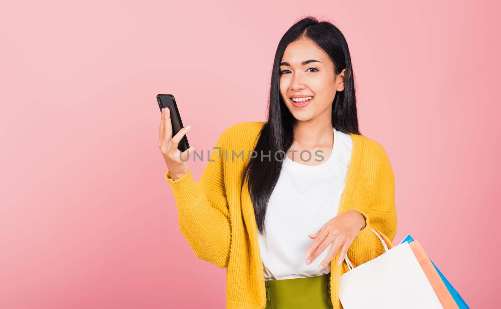 Portrait Asian happy beautiful young woman teen shopper smiling standing excited holding online shopping bags online colorful multicolor and smartphone on hand, studio shot isolated on pink background