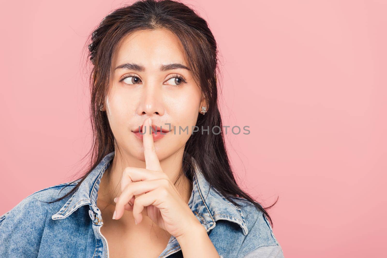 Portrait Asian beautiful young woman wear denim smile stand making finger on lips mouth silent quiet gesture, studio shot isolated on pink background, Thai female quiet hand sign copy space