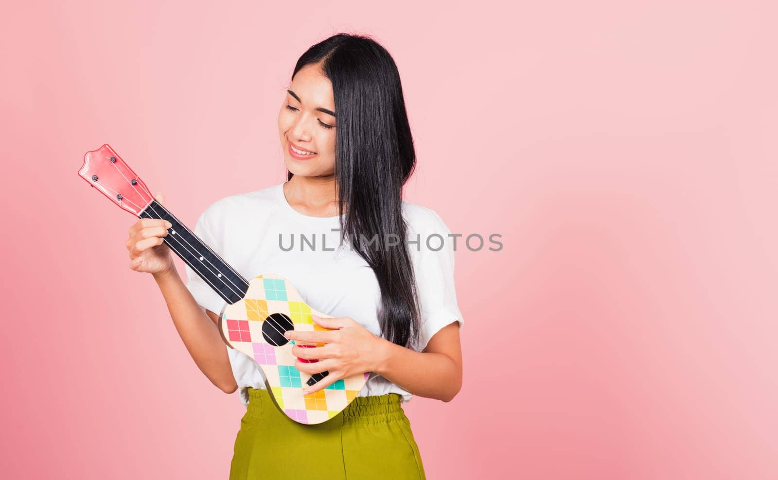 Portrait of happy Asian beautiful young woman teen confident smiling face hold acoustic Ukulele guitar, female playing Hawaiian small guitar, studio shot isolated on pink background, with copy space