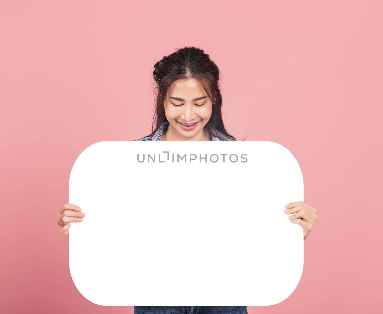 Happy Asian beautiful young woman smiling excited wear denims holding empty speech bubble sign, Portrait female posing show up for your idea looking at bubble, studio shot isolated on pink background
