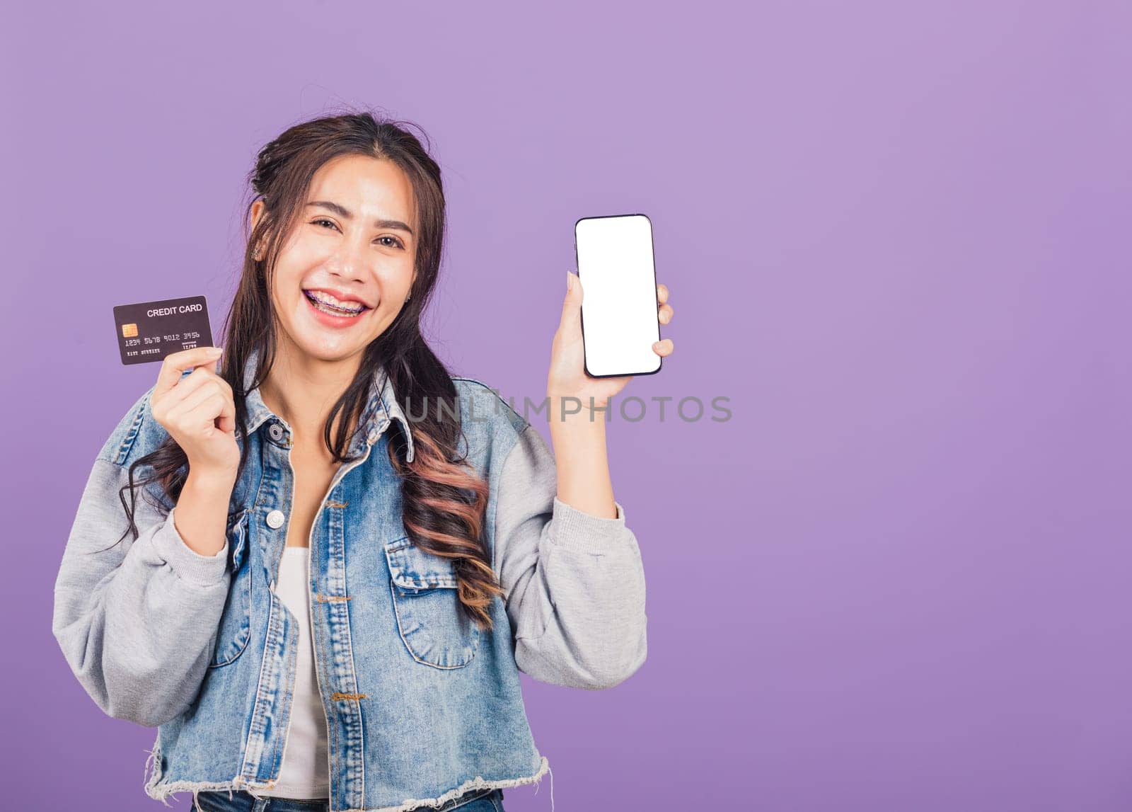 Portrait of Asian beautiful young woman confident wear denim jeans shirt smiling show mobile phone blank screen and credit card bank, studio shot isolated on purple background