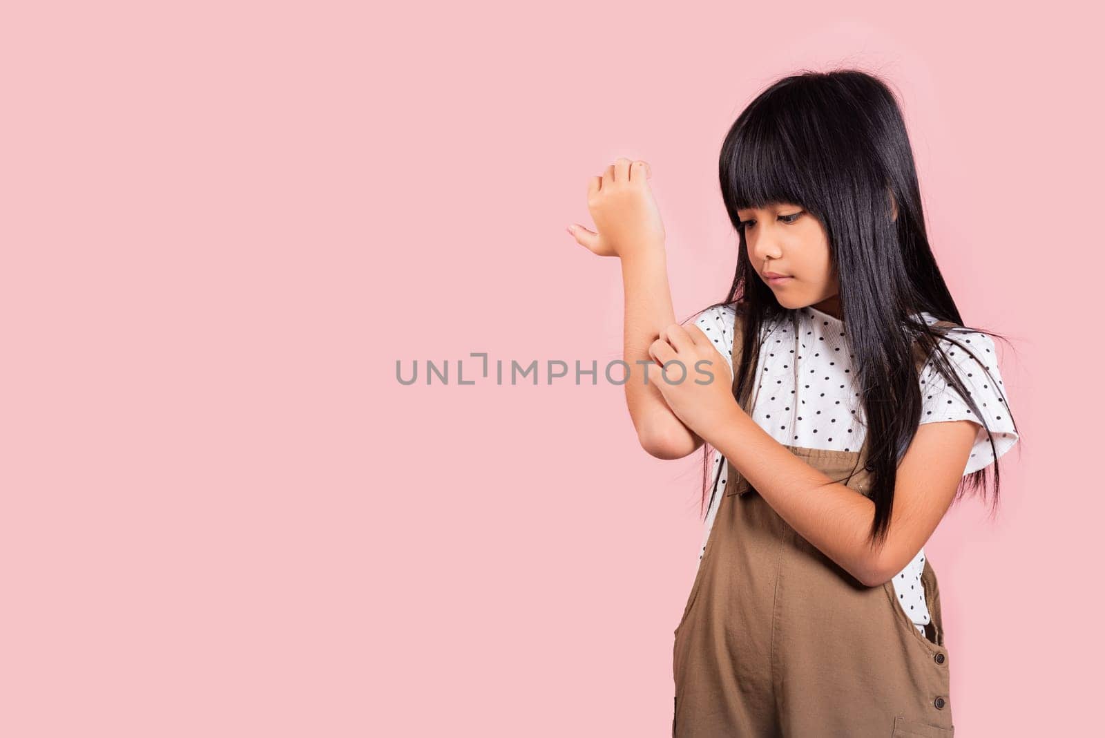 Asian little kid 10 years old scratching itch arm from a mosquito bite at studio shot isolated on pink background, Portrait of Happy child girl dermatitis and scabies, Allergy symptoms, malaria day