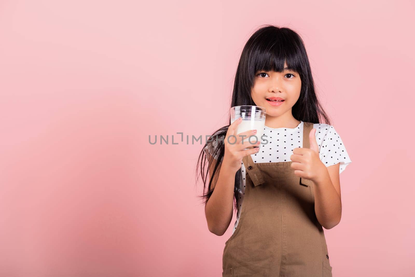 Asian little kid 10 years old smile hold milk glass drink white milk and show thumb up finger for good sign at studio shot isolated on pink background, Happy child girl daily health care medicine food