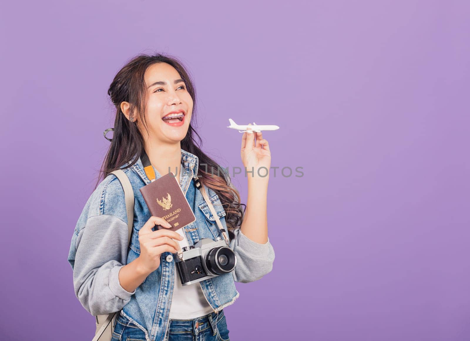 Happy Asian portrait beautiful young woman excited smiling in summer vacation holding airplane toy, passport and vintage photo camera, Thai female ready travel trip isolated on purple background