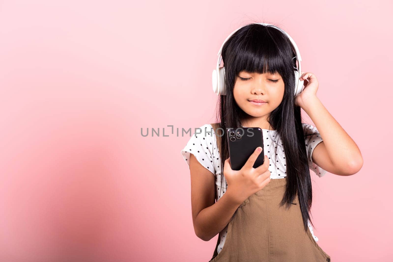 Asian kid 10 years enjoying listening music from mobile phone wear wireless headset closed eyes at studio shot isolated on pink background, Happy child girl use smartphone listen music with headphones