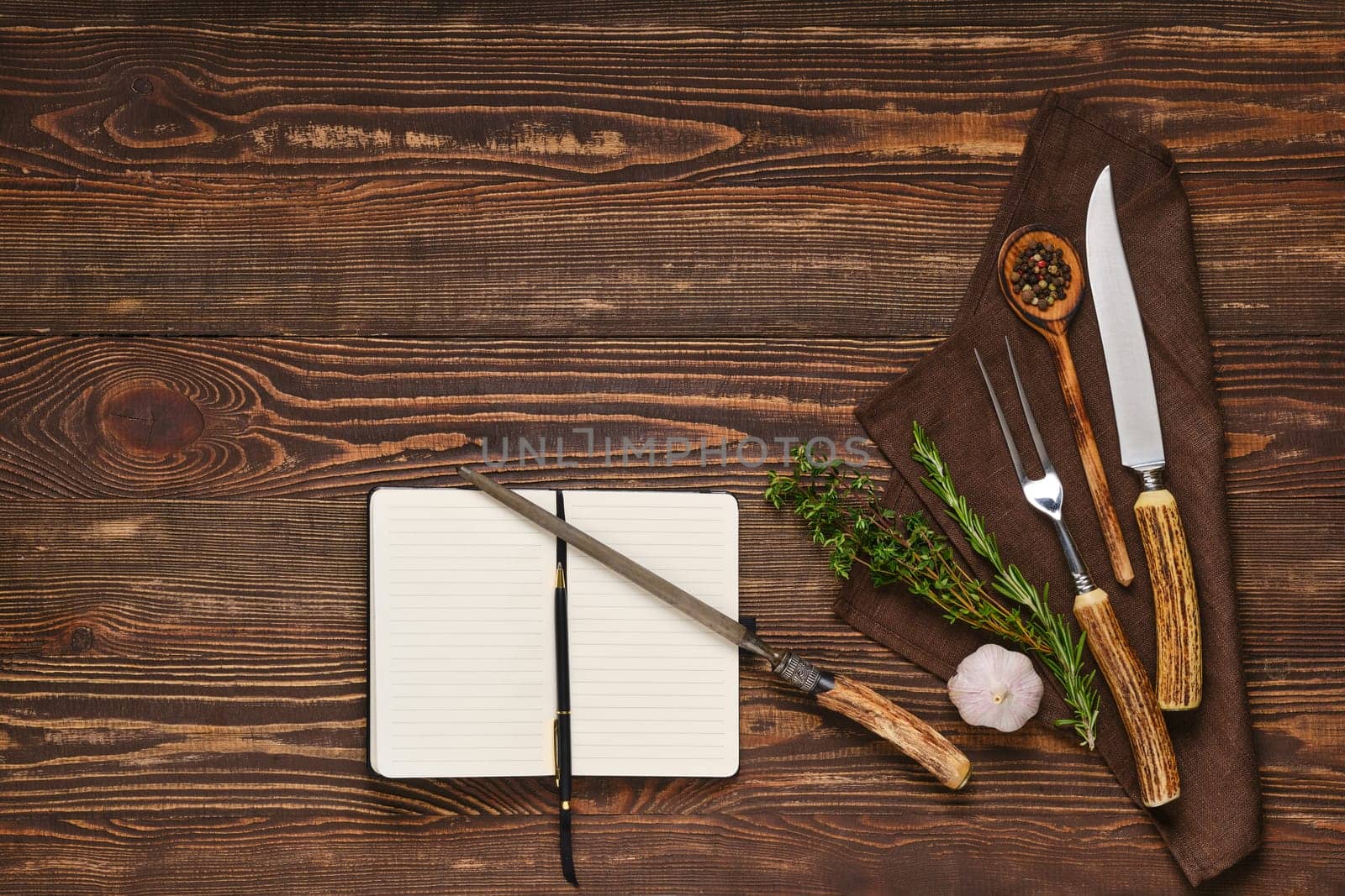 Vintage steak fork, knife and sharpener on wooden table next to notebook. Barbecue appliances and open notepad, food blogging and menu concept.