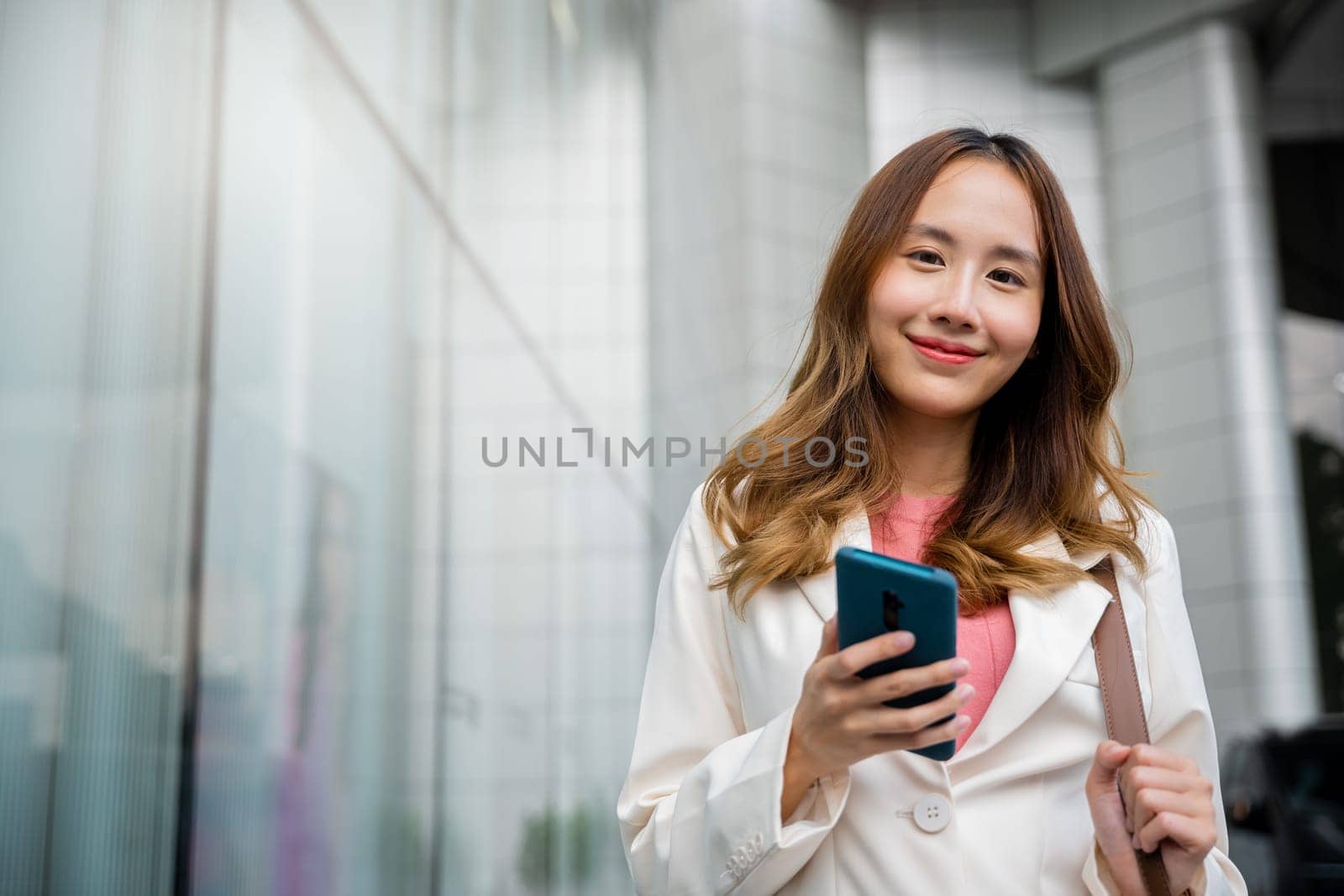 Asian businesswoman texting smartphone commuting work she walking near her office building, Happy business woman smiling using mobile phone outdoor walking on city street urban looking to camera