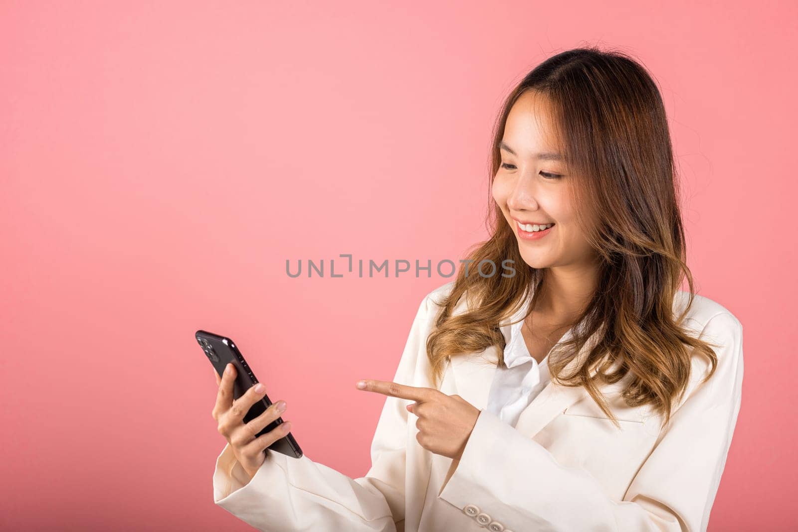 Portrait Asian happy beautiful young woman smile making finger pointing on screen smartphone in hand shopping online application looking to screen, studio shot isolated pink background with copy space
