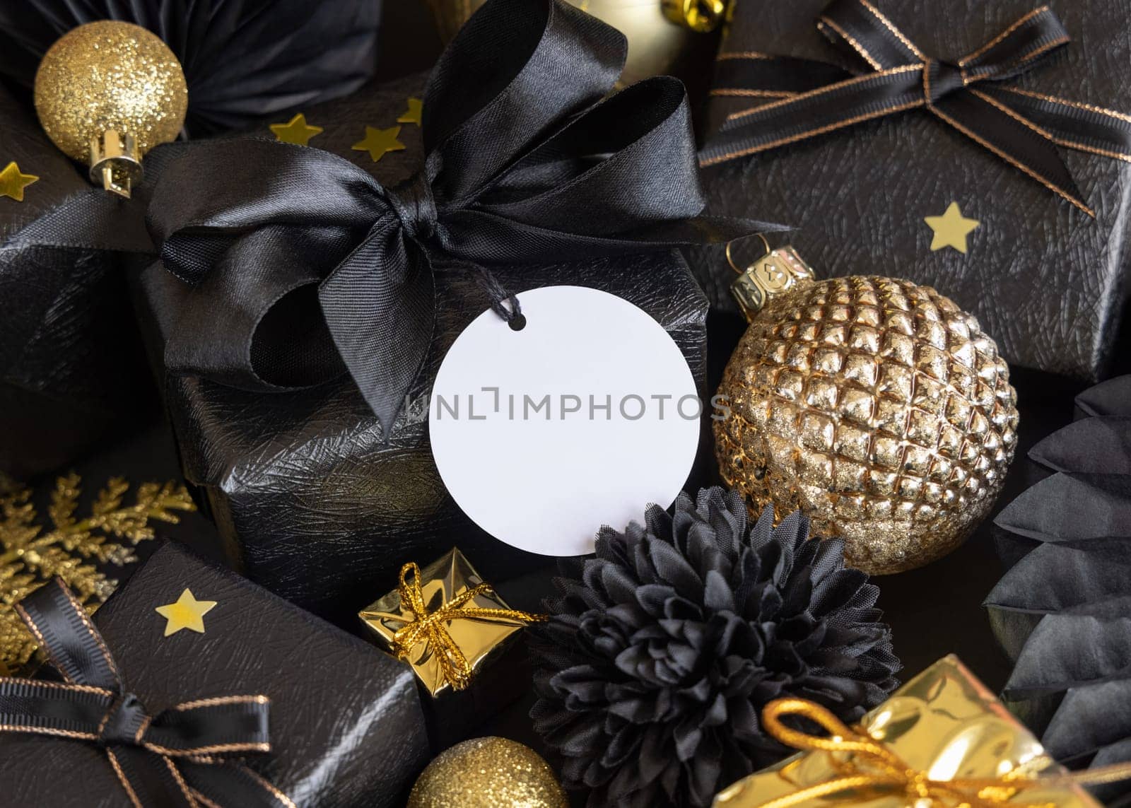 Black and golden Christmas Gift Boxes with a bow and ornaments close up, round paper gift tag mockup, copy space. Dark winter composition with blank label card for Christmas, New Year, Birthday, Anniversary