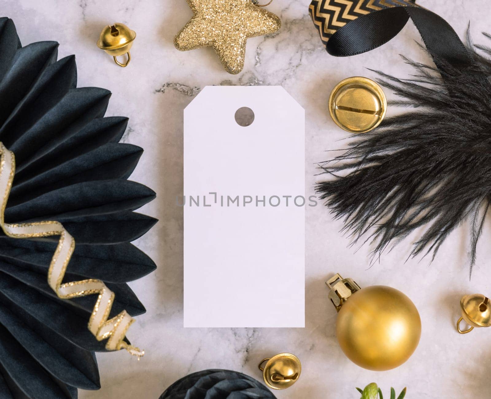 Paper vertical gift tag between fir tree branches and black and golden Christmas decorations on a white marble table top view. Mockup, copy space. Winter holiday flat lay with blank label