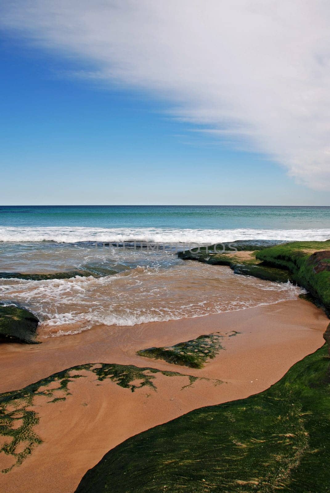 Sandy beach with blue ocean and blue sky and moss covered rocks