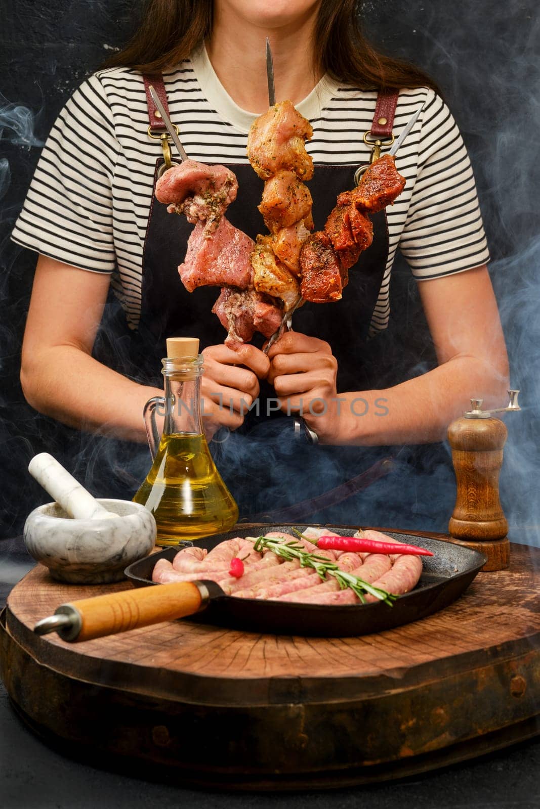 Woman with different meat on skewers stands in front of pan with sausages