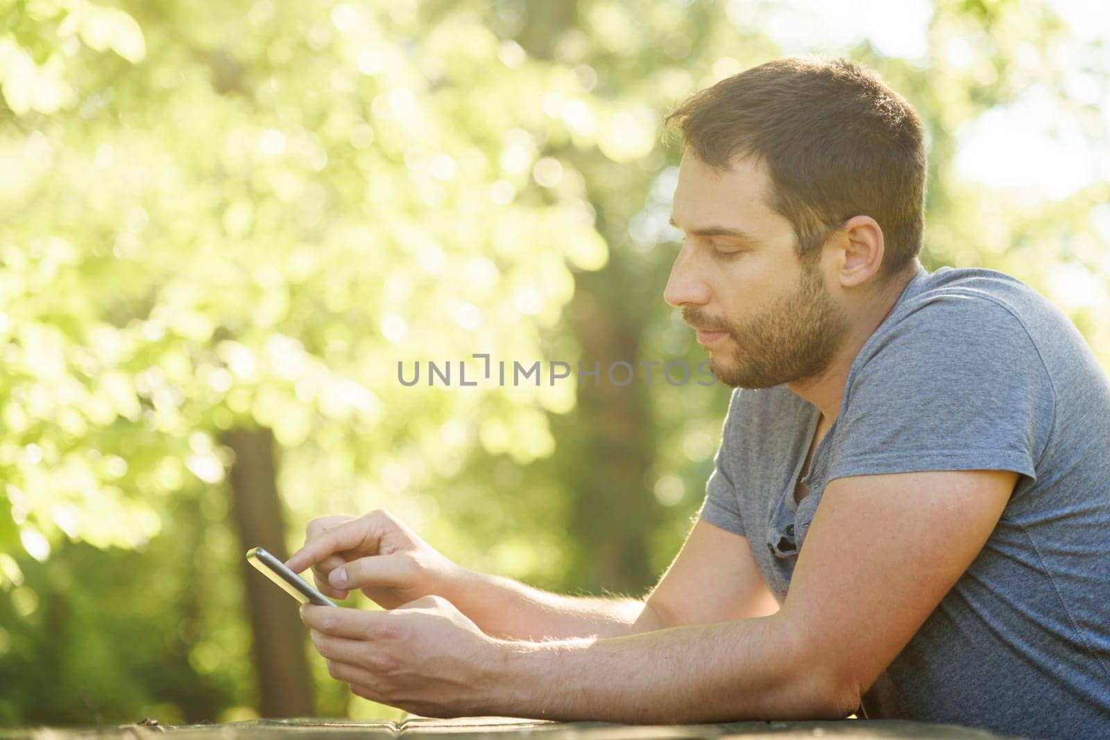 Young man sitting in park, relaxing and using mobile smart phone for social media leisure. Man holding cell phone and texting. Green nature bokeh background with copyspace.