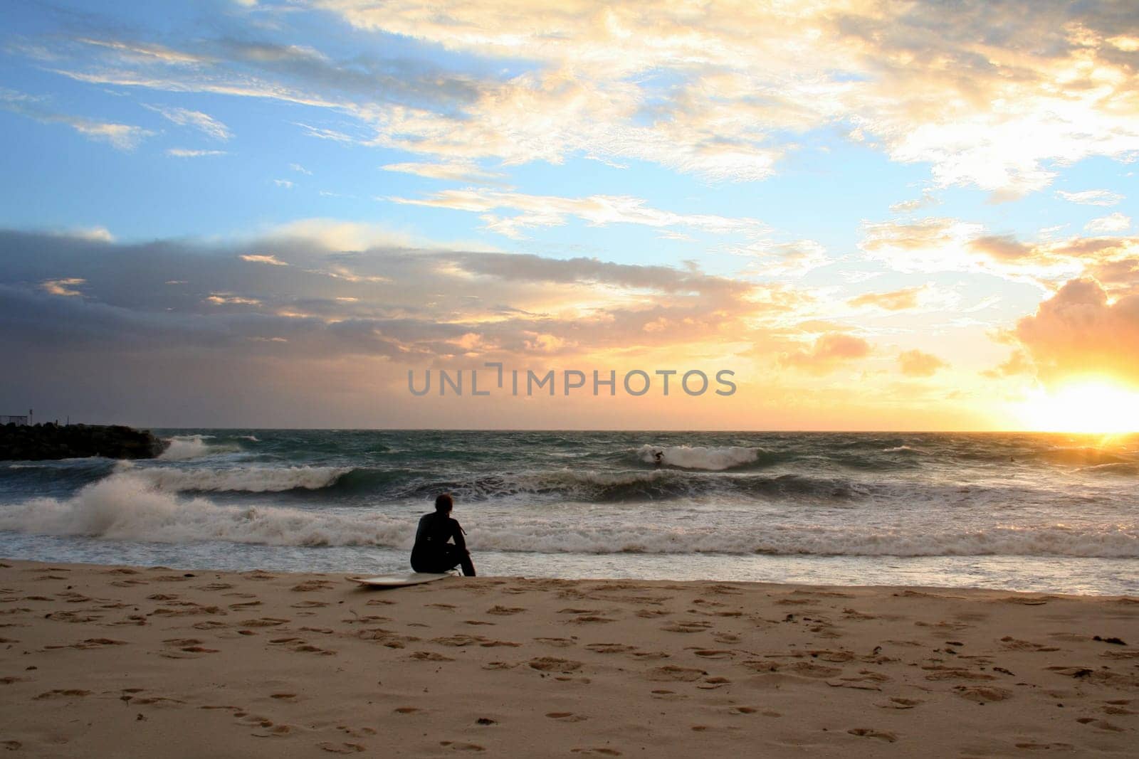 Image of a surfer sitting on his board at the beach, looking over the waves as the sun sets on the horizon.