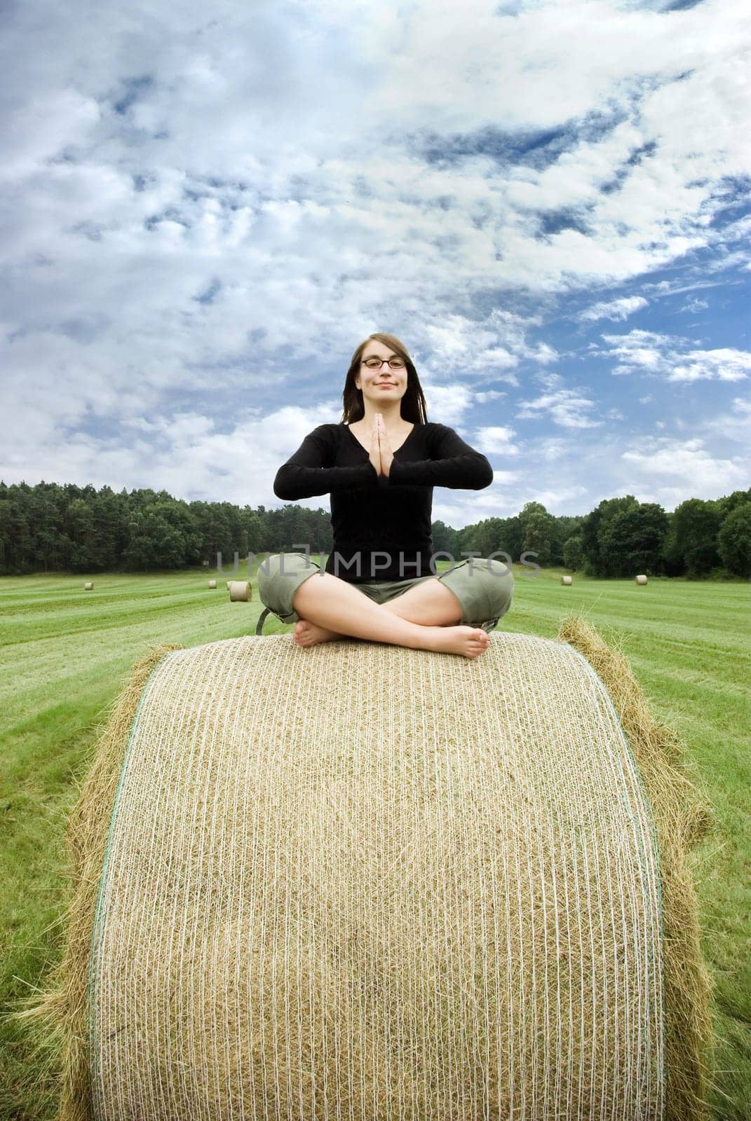 young woman sitting on hay bale meditating in front of a beautiful landscape with blue sky