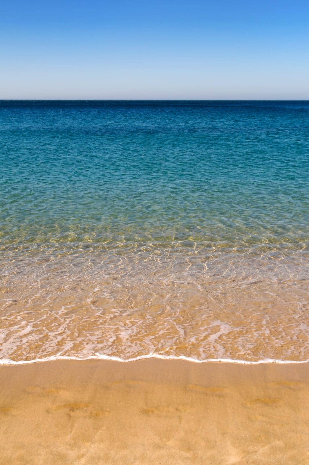 A sandy beach, with water lapping at the shore leading to blue sky