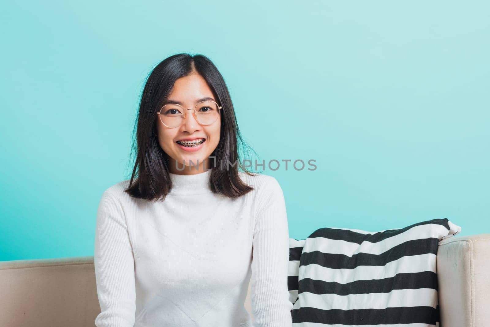Asian beautiful young woman wearing eyeglasses sitting on sofa looking at camera, portrait relaxation of happy female smiling in living studio shot isolated on blue background
