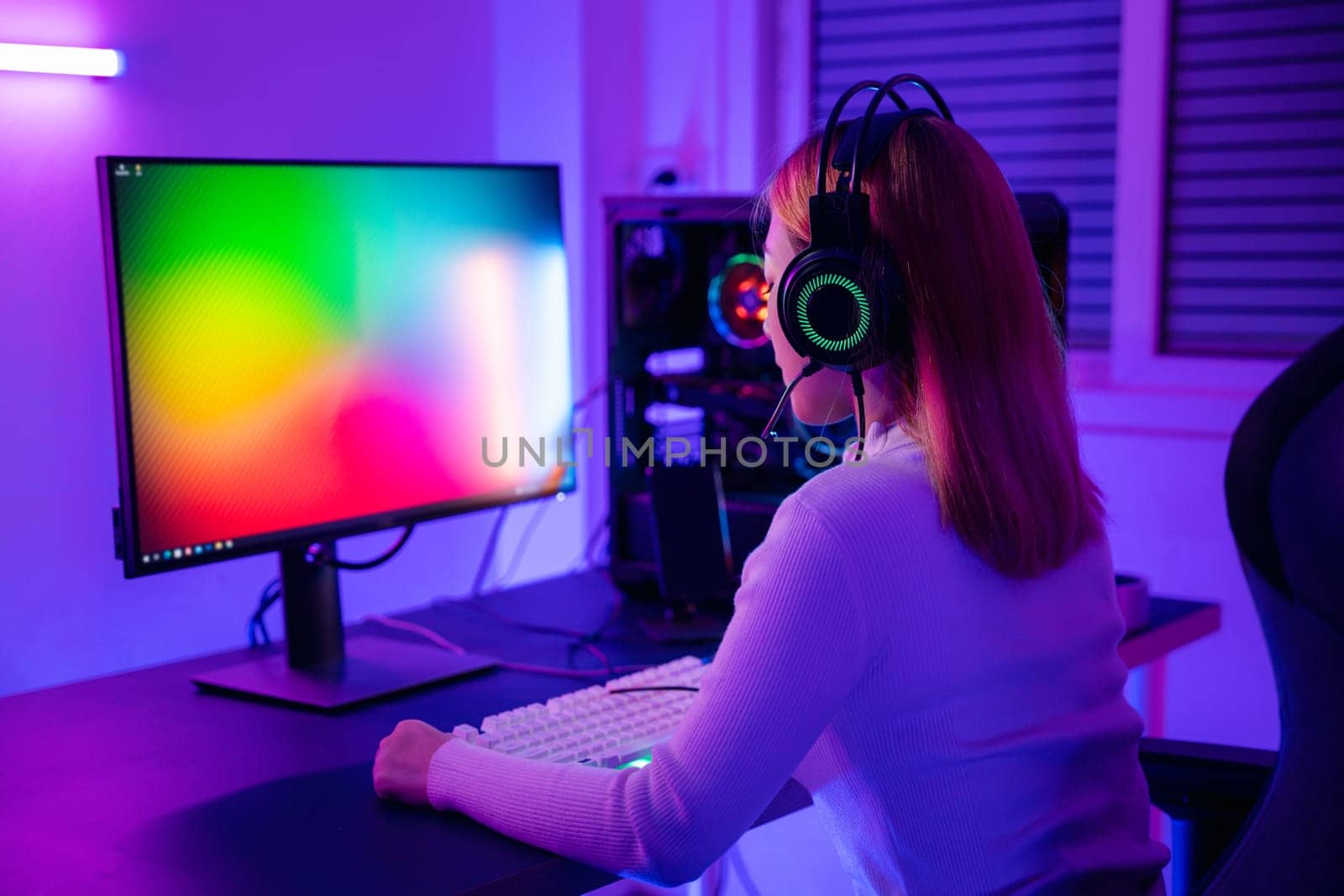 Asian professional gamer playing online video game on desktop computer PC have colorful neon LED lights, young woman in gaming headphones using computer for playing game at home