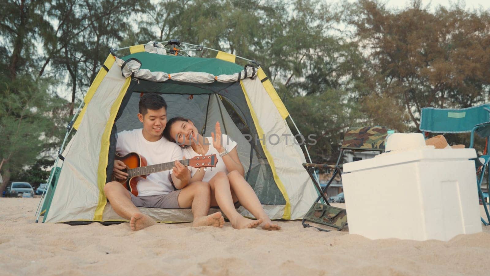 Young loving couple pretty while camp playing guitar while sitting at the camping tent on beach in holiday vacation time at sunset, Young Asian group woman and man in summer travel outdoor