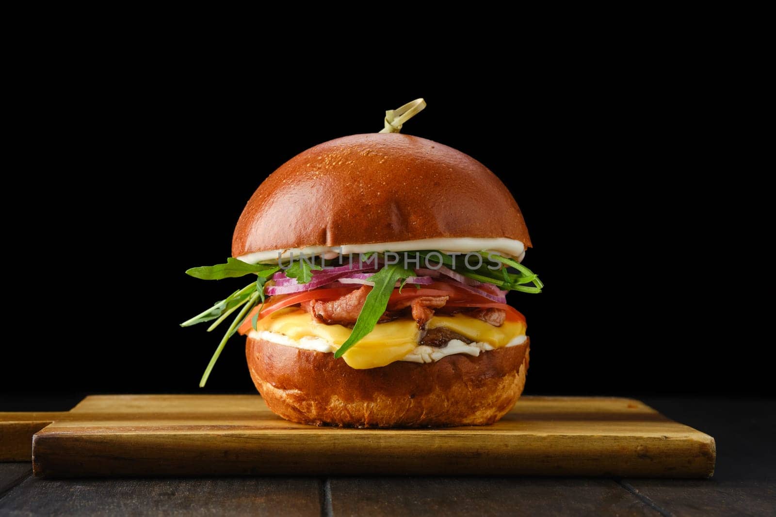 Burger with beef and crispy bacon on wooden serving board over black background