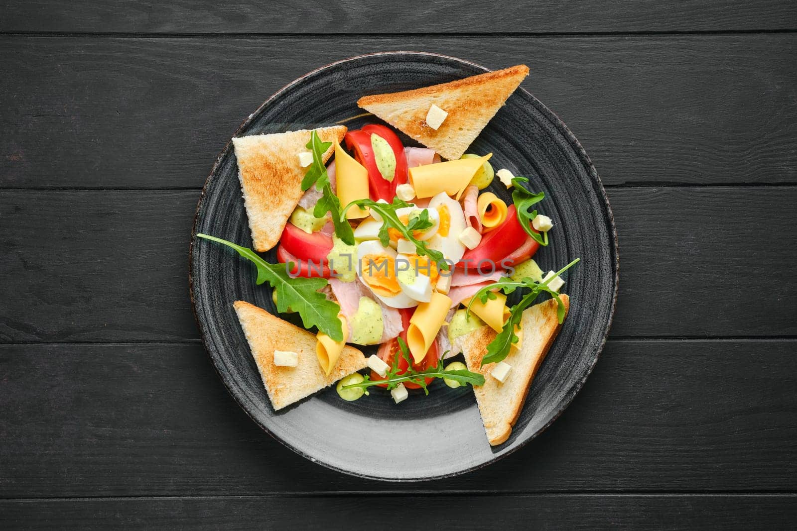 Overhead view of boiled eggs with slices of toast, cheddar, ham and tomato on black wooden table