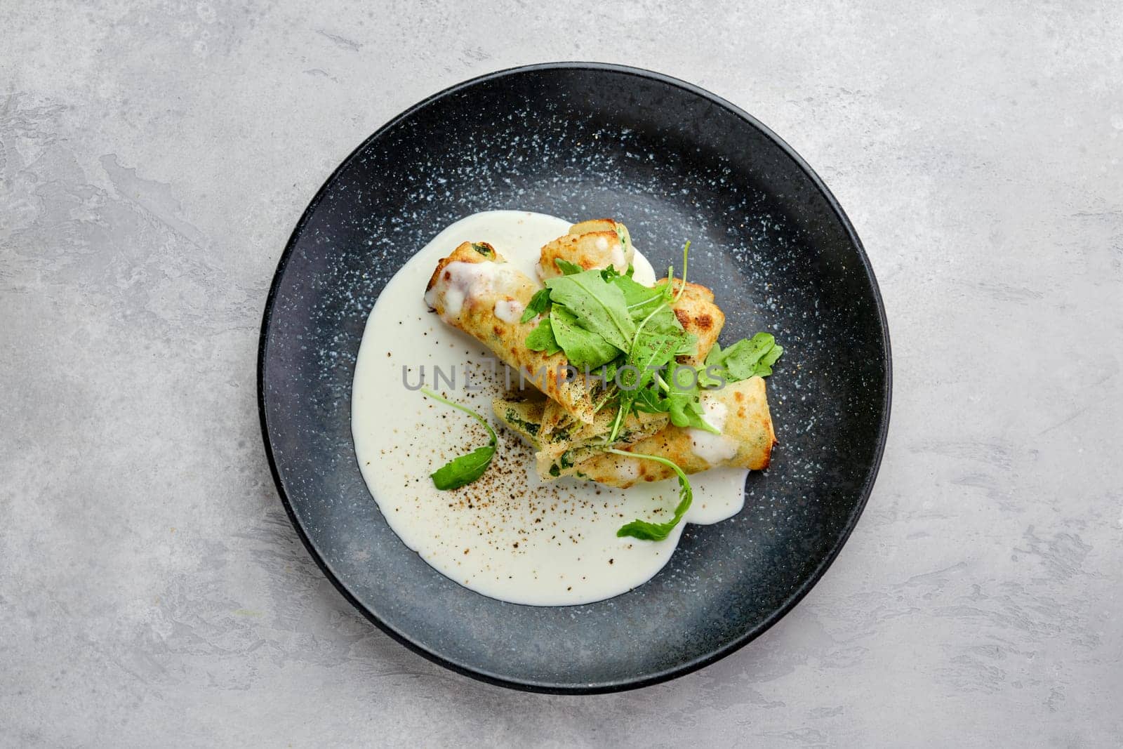Top view of thin crepe stuffed with cheese, arugula and spinach on a plate with creamy sauce