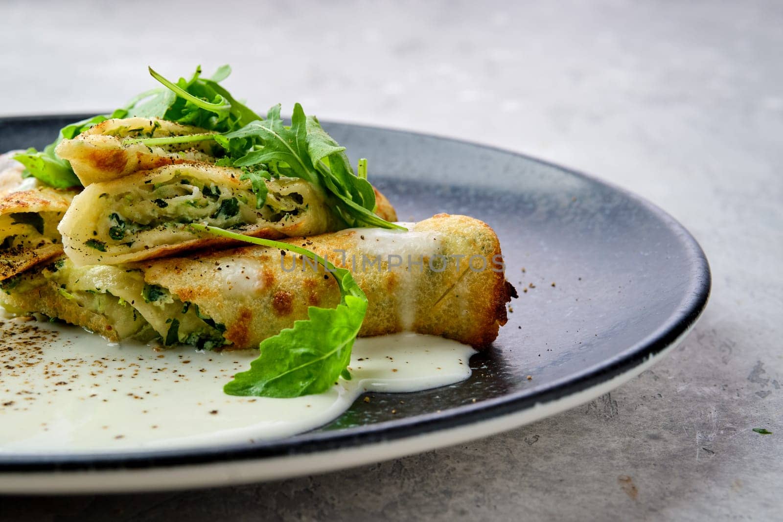 Closeup view of thin crepe stuffed with cheese, arugula and spinach on a plate with creamy sauce