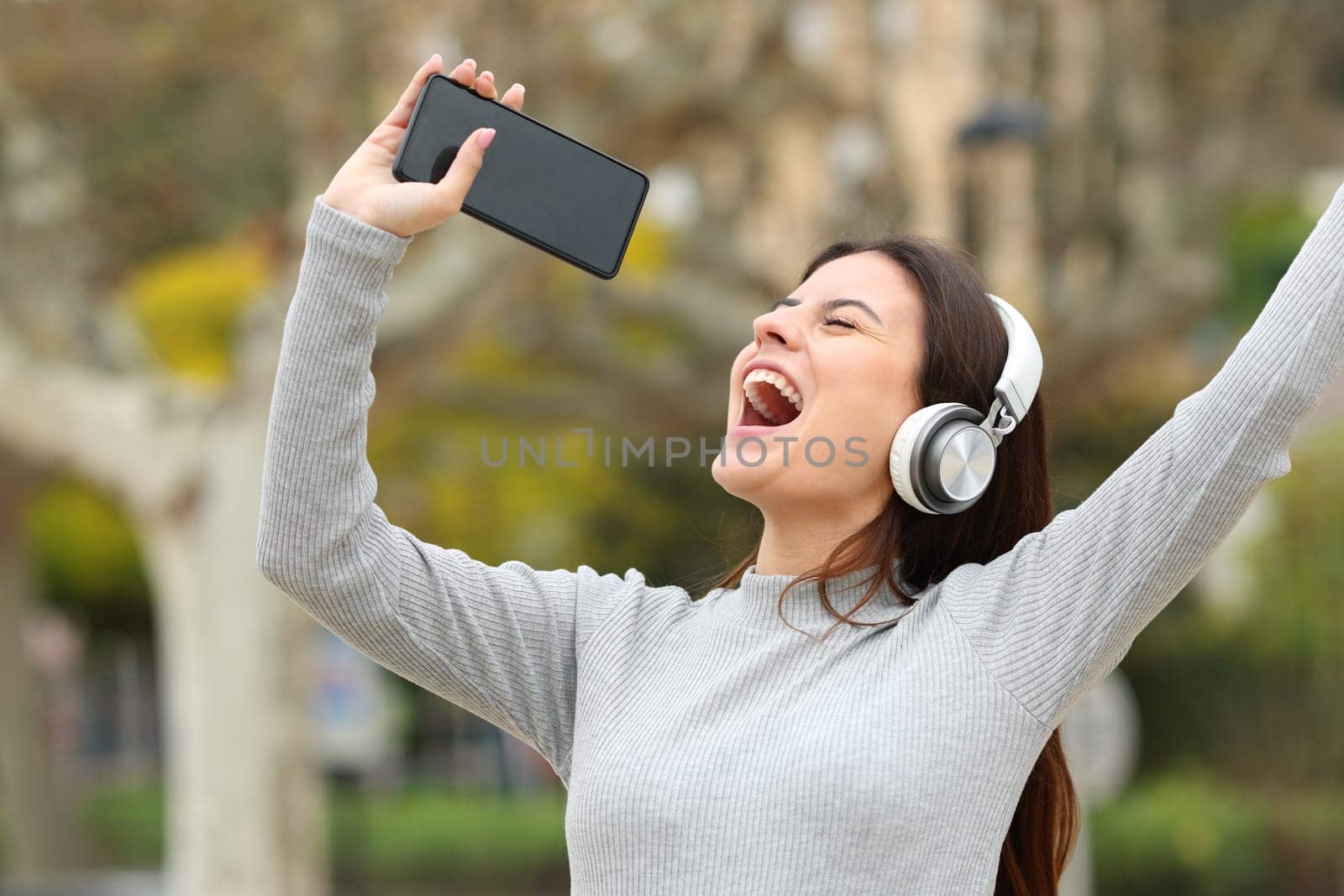 Happy teen singing listening to music showing smart phone screen in a park