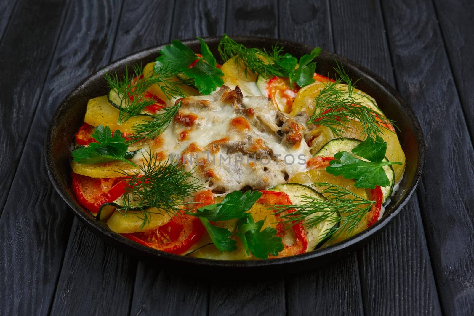 Meat with cheese and slices of potato, tomato and zucchini baked in oven in cast iron pan