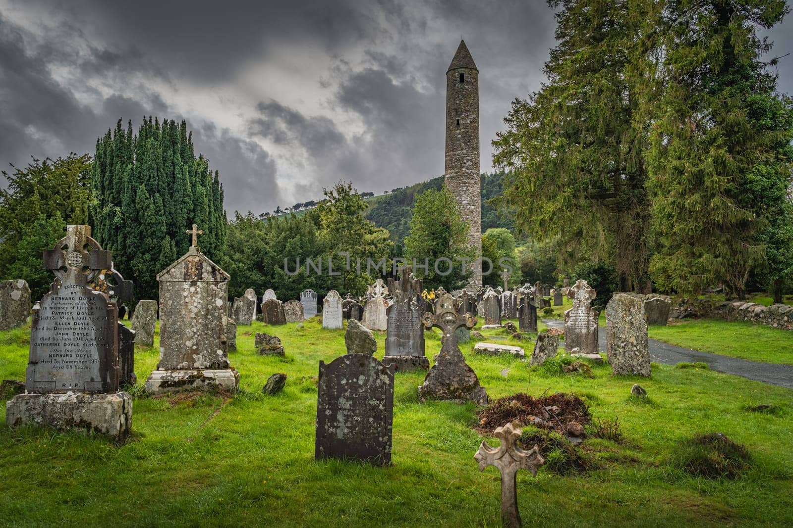 Wicklow, Ireland, August 2019 Medieval tower and ancient graves with Celtic crosses in Glendalough Cemetery. Moody storm sky in background