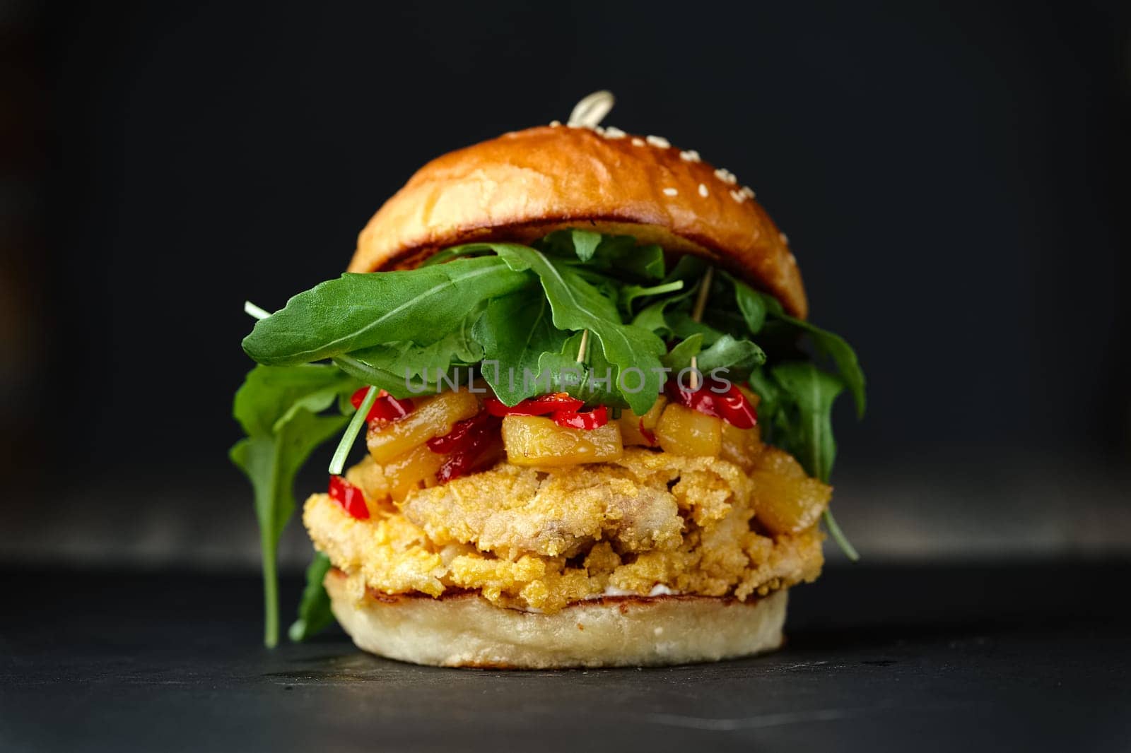Close-up photo if chickenburger on black background with cheese, bacon, tomatos, green salad and chili.