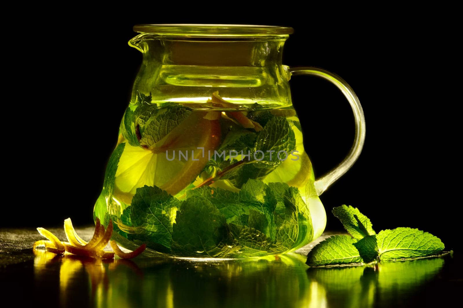 Transparent teapot of green tea with lemon and mint with ingredients. Jar of  green tea with lime, lemon, mint on black slate stone board over black textured background.