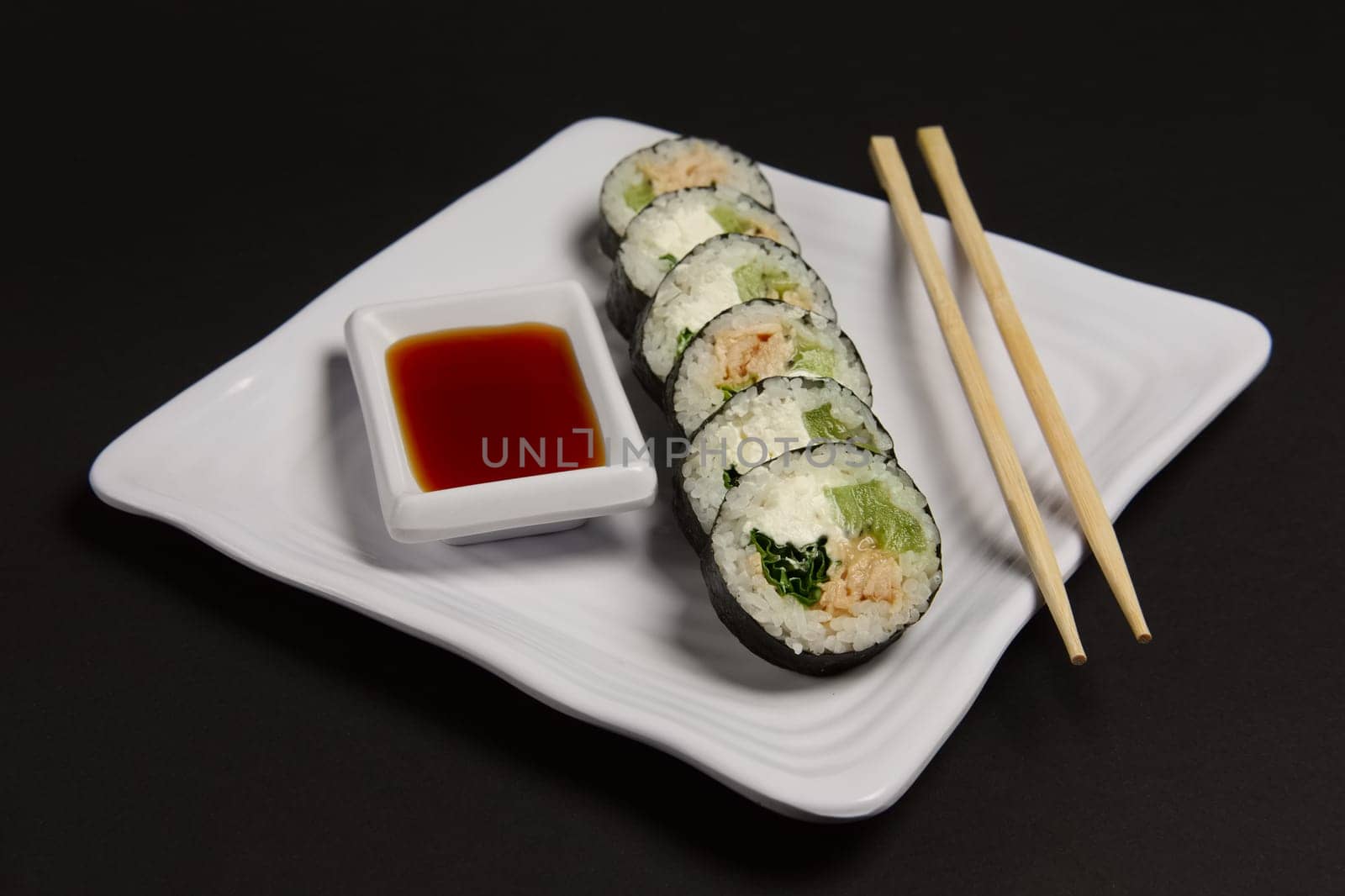 Angle view of portion of chicken teriyaki rolls served on black paper with soy sauce and hashi.