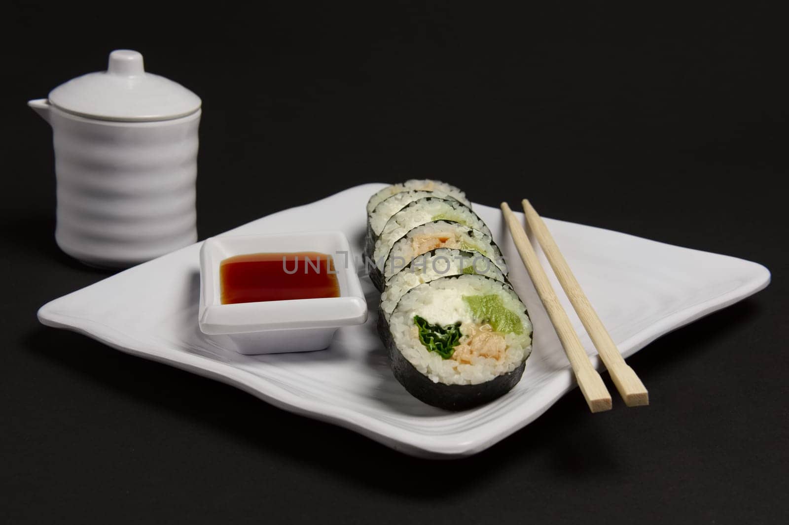 Angle view of portion of chicken teriyaki rolls served on black paper with soy sauce and hashi.