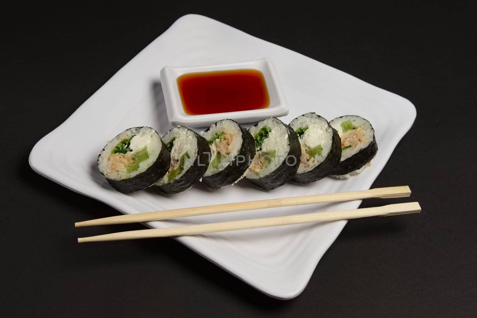 Angle view of portion of chicken teriyaki rolls served on black paper with soy sauce and hashi.