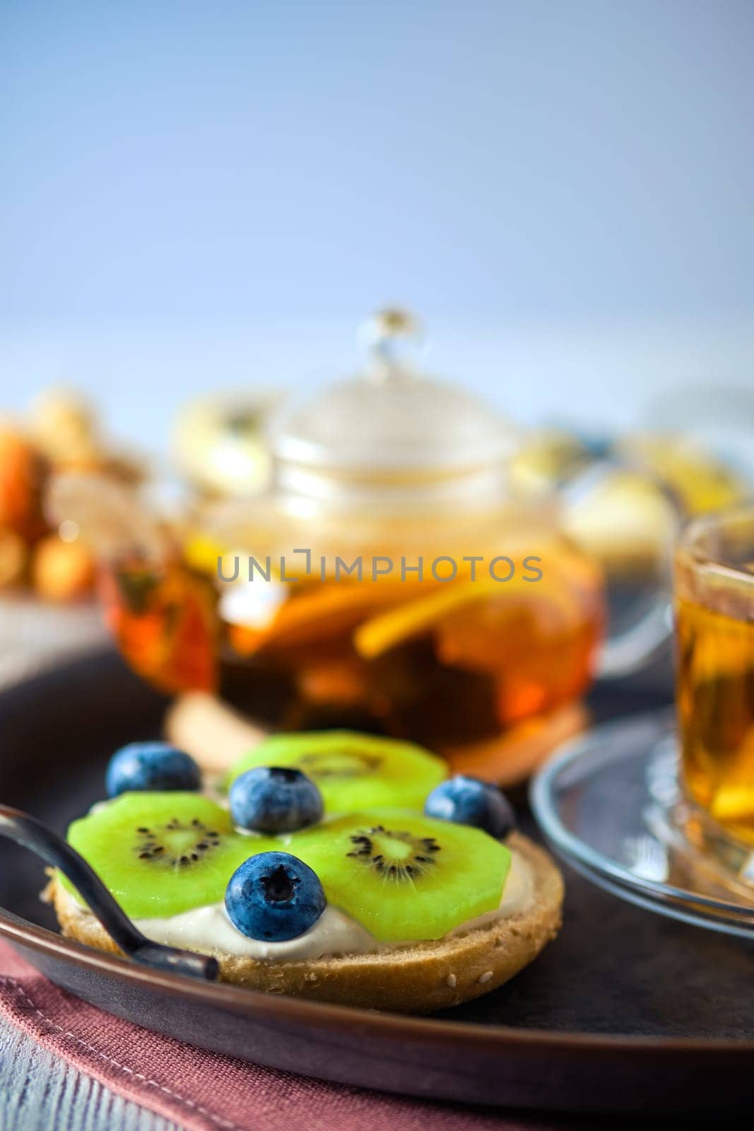 selective focus photo of cottage cheese on fresh bun with kiwi and blueberry and tea on background