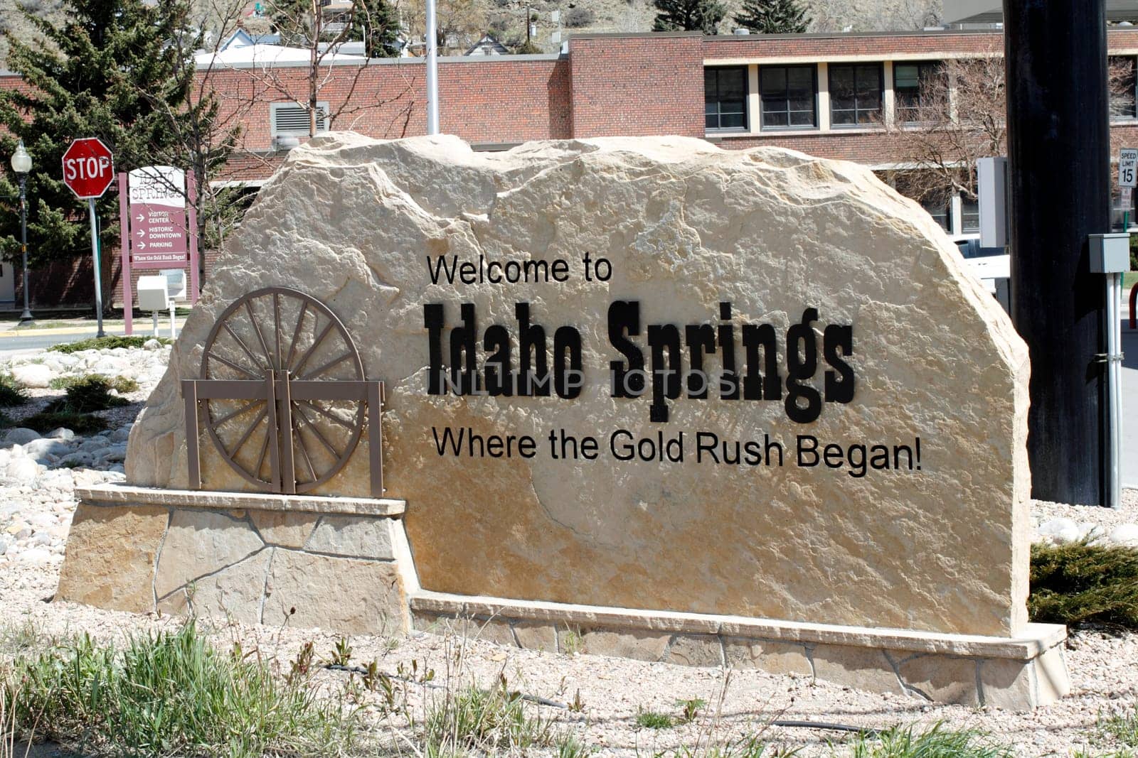 Idaho Springs, CO, USA - April 23, 2014: Large tan stone that has written on and carved into it, Welcome to Idaho Springs, Where the Gold Rush Began! The sign also has a brown sculpture of a water wheel on a sunny day in this small town.