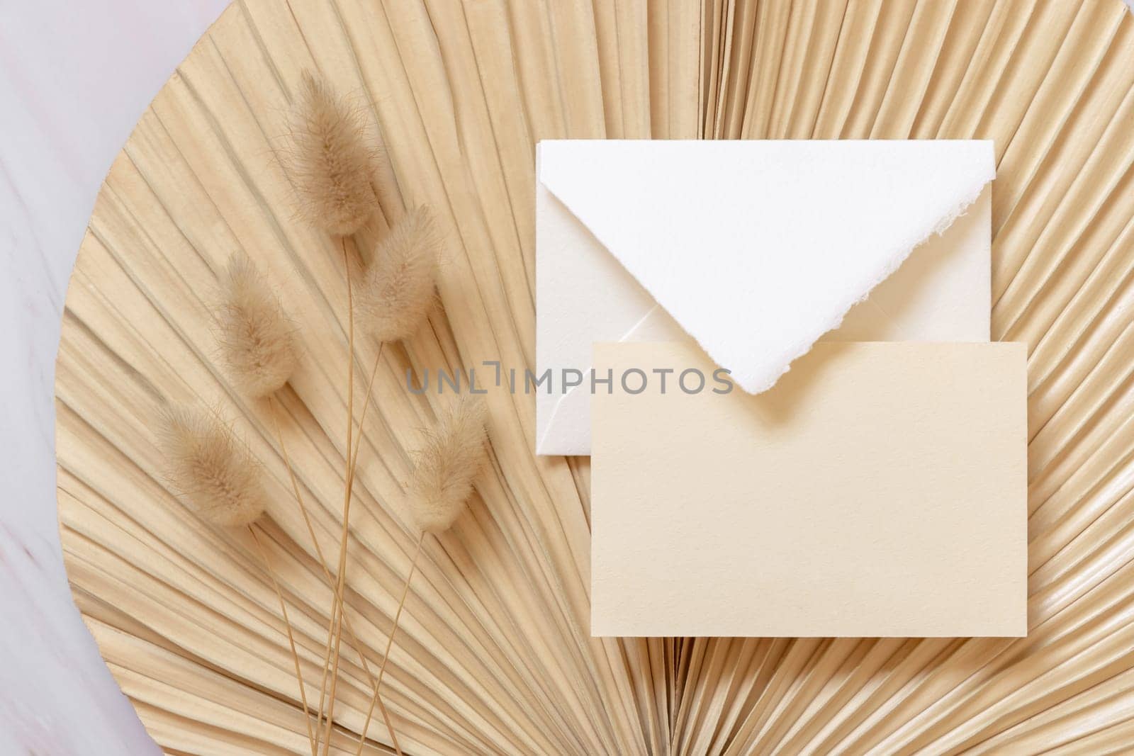 Blank card and envelope on dry beige palm leaf near hare's tail grass top view, mockup. Boho scene with horizontal card. Bohemian or close to nature greeting card or wedding Invitation.