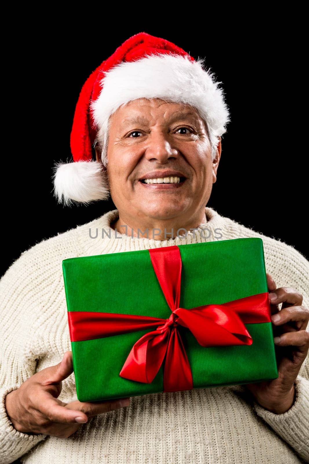 Shyly grinning old gentleman with a red Santa Claus cap and white knitted pullover. He is holding a green wrapped present with both hands in front of his chest. Gift giving theme. Isolated on black.