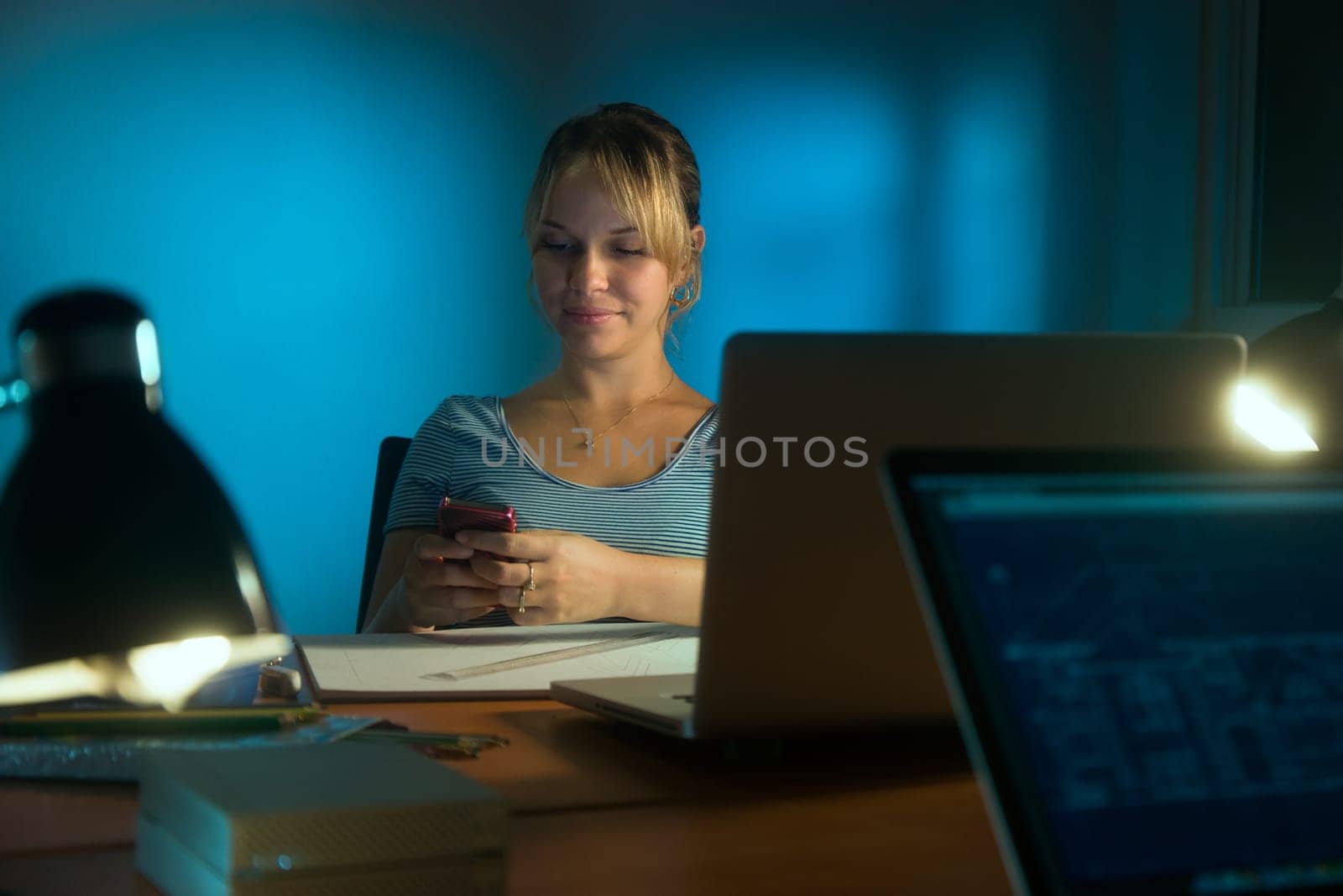 Beautiful woman working as interior designer, staying late at night in office with drawings and laptop computer to complete a project. The girl types a message on mobile phone.