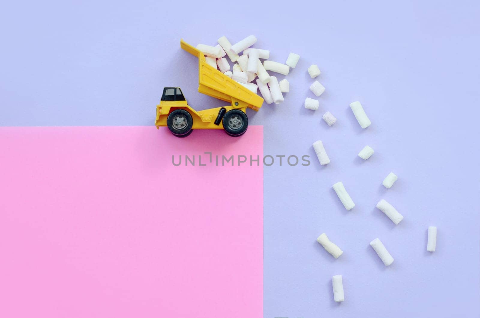 Yellow little toy dump truck throws marshmallow pieces from its raised back on a pastel violet and pink background. Flat lay minimal top view.